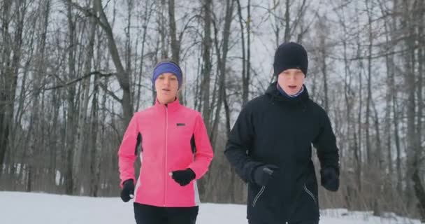 Joven hombre y mujer en una carrera matutina en el bosque de invierno. Una mujer con una chaqueta suelta un hombre con una chaqueta negra corre por un parque de invierno. Vida sana familia feliz . — Vídeos de Stock