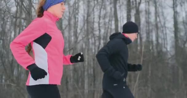 Joven hombre y mujer en una carrera matutina en el bosque de invierno. Una mujer con una chaqueta suelta un hombre con una chaqueta negra corre por un parque de invierno. Vida sana familia feliz . — Vídeos de Stock