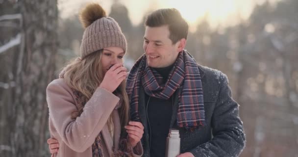 Счастливая пара держит чашки горячего чая над зимним пейзажем. Молодая пара влюблена на зимних каникулах, стоит рядом с деревом и пьет горячий чай — стоковое видео