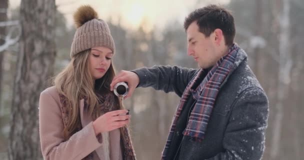 Любящая пара мужчина и женщина в зимнем лесу пьют чай из термоса. Стильные мужчина и женщина в пальто в парке зимой на прогулку. Медленное движение — стоковое видео