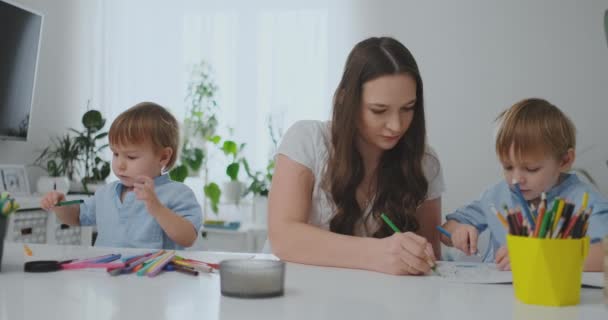 Beyaz bir masada oturan iki çocuk ile genç bir anne yavaş hareket kağıda renkli kalemler çekiyor — Stok video