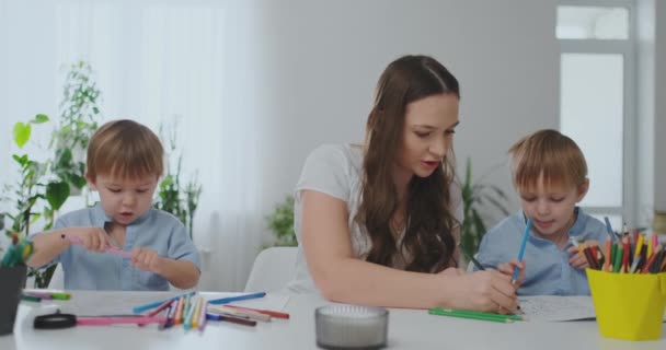 Una familia de dos niños y una madre joven sentada a la mesa dibuja sobre papel con lápices de colores. Desarrollo de la creatividad en los niños. blanco interior limpio — Vídeos de Stock