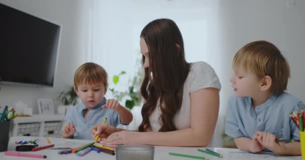 Молода мати з двома дітьми сидить за білим столом малює кольорові олівці на папері, допомагаючи робити домашнє завдання — стокове відео