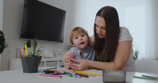 한 어린이 소년과 식탁에 앉아 있는 한 젊은 어머니가 한 가족은 색연필로 종이를 그립니다. 어린이 창의력 개발. — 비디오