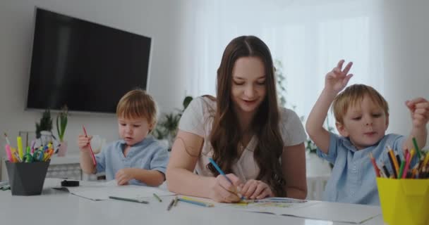 Μια νεαρή μητέρα με δύο παιδιά που κάθονται σε ένα λευκό τραπέζι ζωγραφίζει χρωματιστά μολύβια σε χαρτί βοηθώντας να κάνουν την εργασία — Αρχείο Βίντεο
