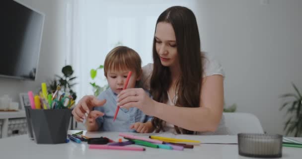 Am Wohnzimmertisch sitzend, bringt eine junge Mutter ihrem Sohn das Halten eines Bleistifts bei und bringt ihm das Zeichnen bei — Stockvideo