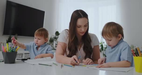 En ung mor med två barn sitter vid ett vitt bord ritar färgade pennor på papper i slow motion — Stockvideo