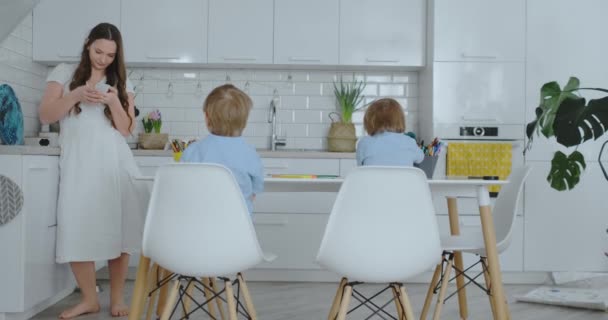 Masada oturan mavi gömleklere iki oğul ve beyaz bir masada oturan kağıt üzerinde kalemler çizmek, ve annem Smartphone bir metin mesajı yazar — Stok video