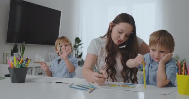 Mladá matka se dvěma dětmi, hovořije na mobilním telefonu, kreslí tužkou a pomáhá dětem kreslit barevnými tužkami — Stock video
