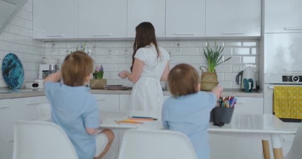 Maman dans la cuisine lave la vaisselle et deux fils assis à un bureau dessin sur papier avec des crayons de couleur. Jeune famille maman sourit et regarde les enfants — Video