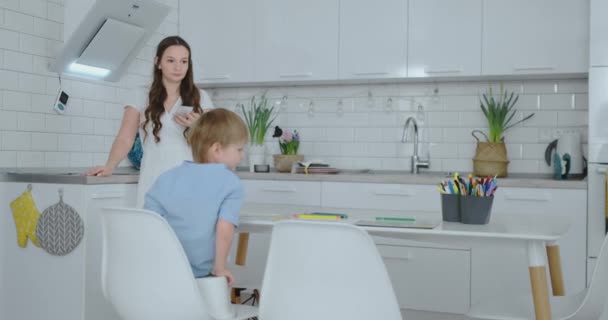 Mãe na cozinha lavar pratos e dois filhos sentados em uma mesa de desenho em papel com lápis de cor. Jovem família mãe sorri e olha para as crianças — Vídeo de Stock