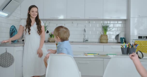 Dois meninos sentados na luz da cozinha desenhar desenhos lápis para missões escolares. Mãe olha para as crianças e sorri na cozinha — Vídeo de Stock