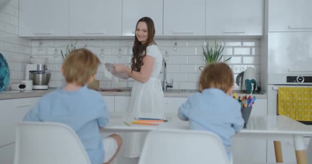 Mãe na cozinha lavar pratos e dois filhos sentados em uma mesa de desenho em papel com lápis de cor . — Vídeo de Stock