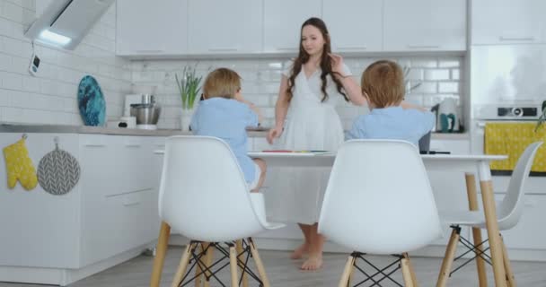 Two boy sitting on kitchen light draw pencils drawings for school-quests. Mom looks at the children and smiles in the kitchen — Stock Video