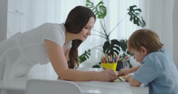 Mamá ayuda a su hijo a realizar la tarea preescolar para dibujar un dibujo a lápiz sentado en la mesa de la casa — Vídeo de stock