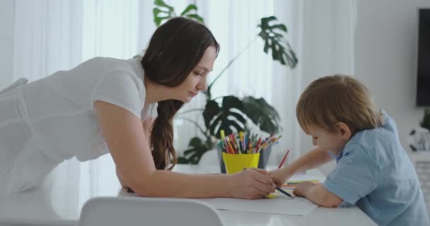 Jonge mooie moeder en zoon tekent met kleurpotloden zitten aan de tafel in de keuken. De camera beweegt in slow motion — Stockvideo