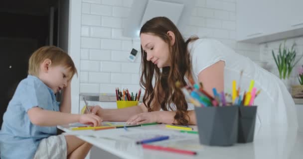 Mamma aiuta il figlio a eseguire i compiti di scuola materna per disegnare un disegno a matita seduto a tavola in casa — Video Stock