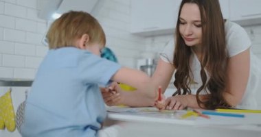 Anne iki oğlu evde masada oturan bir kalem çizim çizmek için okul öncesi ödev gerçekleştirmek için yardımcı olur.