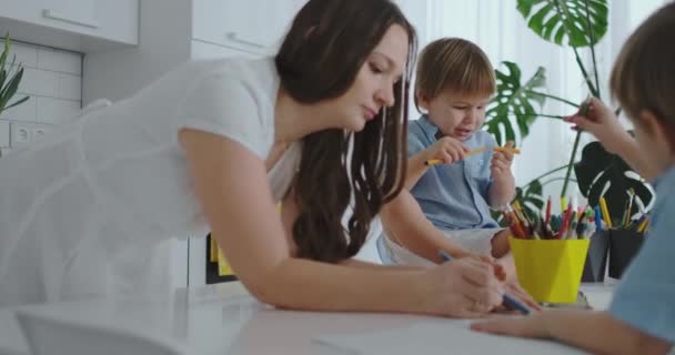 Mamá ayuda a los hijos a aprender a dibujar haciendo tareas de preparación preescolar en casa sentado en la cocina blanca. Dos hermanos niños dibujan un retrato de la madre juntos — Vídeos de Stock