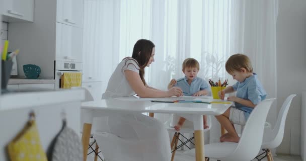 Mom helps sons learn to draw doing homework preschool preparation at home sitting in the white kitchen. — Stock Video