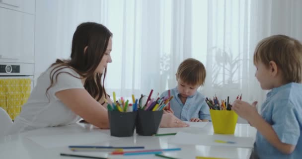 Mamá ayuda a los hijos a aprender a dibujar haciendo tareas de preparación preescolar en casa sentado en la cocina blanca . — Vídeos de Stock