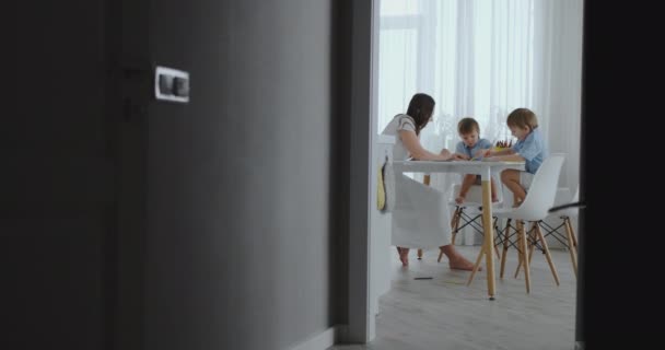 Мама помогает сыновьям научиться рисовать домашнее задание дошкольного образования дома, сидя на белой кухне. Два ребенка братья рисуют портрет матери вместе — стоковое видео