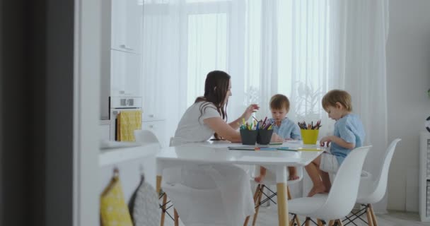 Mamá ayuda a los hijos a aprender a dibujar haciendo tareas de preparación preescolar en casa sentado en la cocina blanca. Dos hermanos dibujan un retrato de mamá juntos — Vídeo de stock