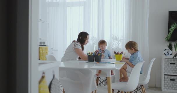 Due figli di ragazzi disegnano con la madre seduta in cucina. Famiglia felice a casa . — Video Stock