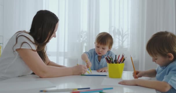 Dos hijos de niños dibujan con su madre sentada en la cocina. Familia feliz en casa. Los hermanos dibujan en la mesa. Gente real . — Vídeo de stock