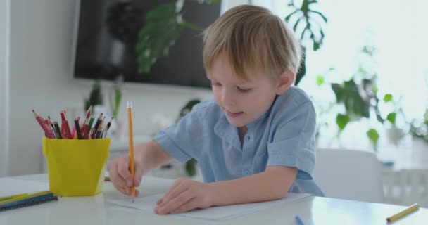 Un niño pequeño sentado en la mesa dibuja un dibujo a lápiz en diferentes colores — Vídeos de Stock