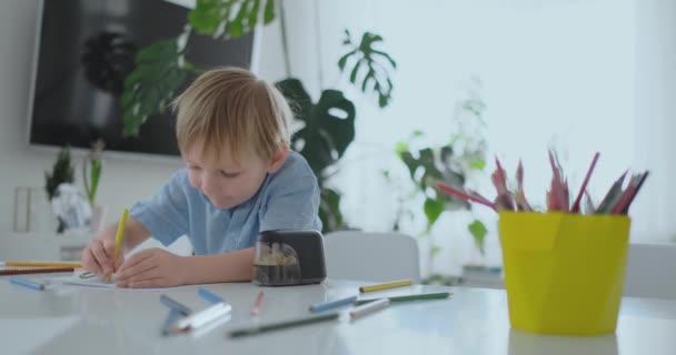 O menino sentado à mesa na sala de estar escreve uma carta para seu pai e desenha uma imagem da família com um lápis — Vídeo de Stock