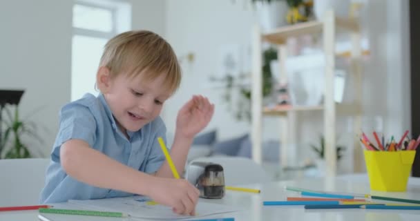 Un niño pequeño sentado en la mesa dibuja un dibujo a lápiz en diferentes colores — Vídeo de stock