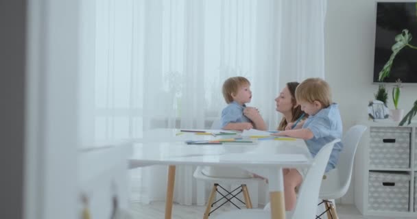 Deux frères garçons s'assoient à une table dans le salon et dessinent avec des crayons sur papier. Maman se lève et étreint les garçons, souriant et caressant sa tête. Louange et aide à faire la tâche — Video