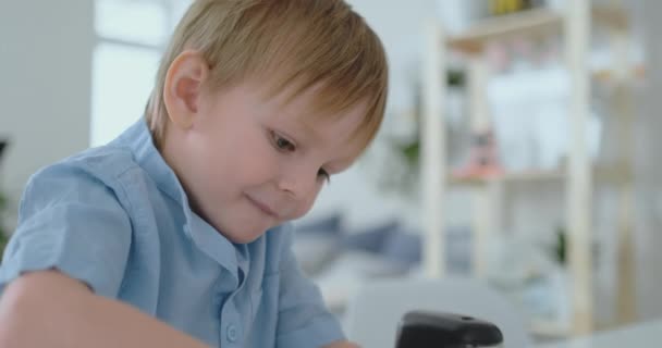 O menino sentado à mesa na sala de estar escreve uma carta para seu pai e desenha uma imagem da família com um lápis — Vídeo de Stock