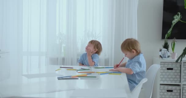 Dois meninos irmãos sentam-se em uma mesa na sala de estar e desenhar com lápis no papel. Mamãe vem e abraça os meninos, sorrindo e acariciando a cabeça dele. Louvores e ajuda a fazer a tarefa — Vídeo de Stock