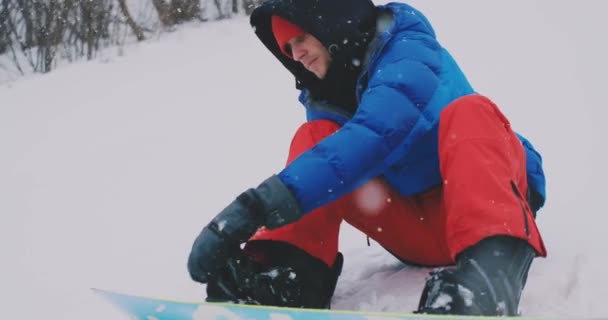 Een man in rode broek zittend op de sneeuw vastmaakt snowboard schoenen en een blauwe jas op de skipiste — Stockvideo