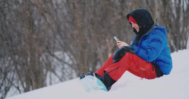 Чоловік сноубордист, який сидить на снігу, фотографує по телефону красивий курортний ландшафт для соціальних мереж. Курортний блогер — стокове відео