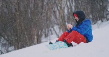 Kar üzerinde oturan bir erkek snowboard fotoğraf bir güzel tatil manzara sosyal ağlar için telefon alır. Blogger Resort. Arkadaşlar smartphone kullanarak metin mesajları yazmak