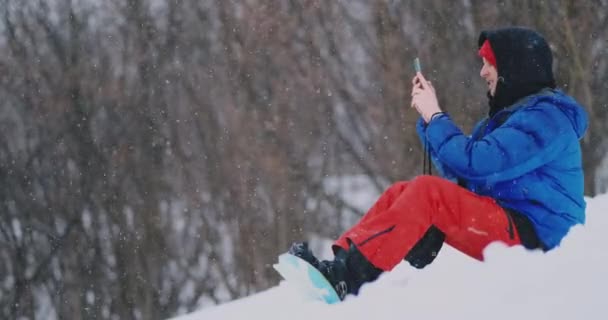 Чоловік сноубордист сидить на снігу фотографує по телефону красиві пейзажі курорту для соціальних мереж — стокове відео