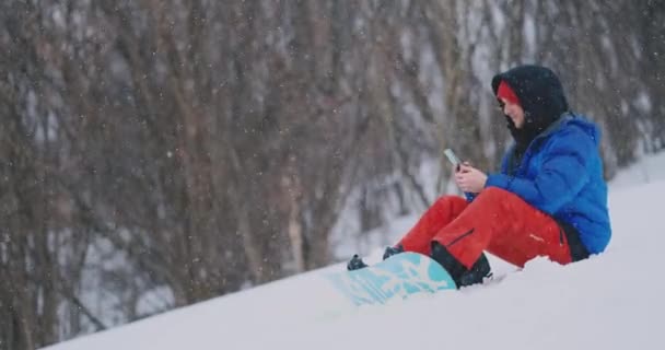 Um snowboarder masculino sentado na neve tira fotos no telefone de uma bela paisagem resort para redes sociais. Um blogueiro no resort. Escreva mensagens de texto para amigos usando seu smartphone — Vídeo de Stock