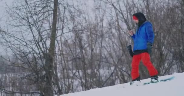 Maschio snowboarder cavalca la tavola sulla pista da sci alla neve e scrivere messaggi per smartphone ai tuoi amici — Video Stock