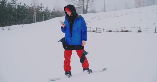 Чоловік сноубордист їде на дошці на лижах до снігового схилу і пише повідомлення на смартфон своїм друзям — стокове відео