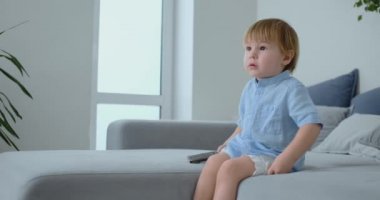 2 yaşında erkek bir koltukta oturur ve onun elinde bir uzaktan kumanda ile oturup Tv izliyor.