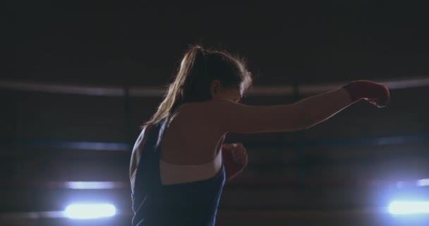 Une belle boxeuse professionnelle se lance dans un combat d'ombre dans une salle sombre au ralenti en vêtements bleus et bandages rouges sur ses poignets. steadicam shot — Video