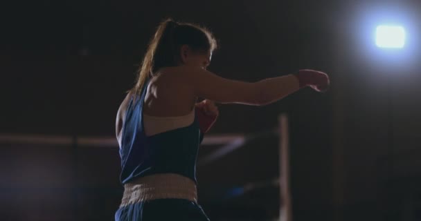 Красивая женщина ведет теневой бой, отрабатывая технику и скорость ударов, усердно тренируясь для будущих побед. Тёмный спортзал. steadicam shot — стоковое видео