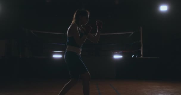 Une belle boxeuse professionnelle se lance dans un combat d'ombre dans une salle sombre au ralenti en vêtements bleus et bandages rouges sur ses poignets. steadicam shot — Video