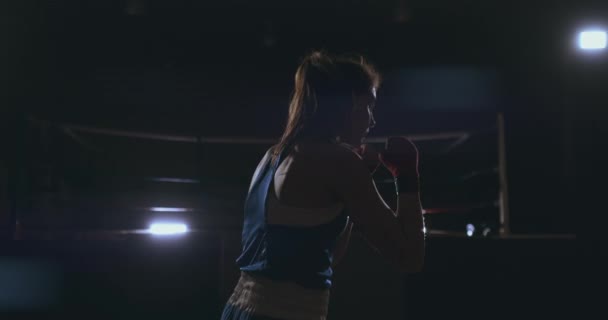 Профессиональная красавица-боксер отбриватыват ударами проводит схватку с тенью в темном зале зала в замедленной съемке в синей одежде и красных бинтах на запястьях. летающие вокруг Земли — стоковое видео
