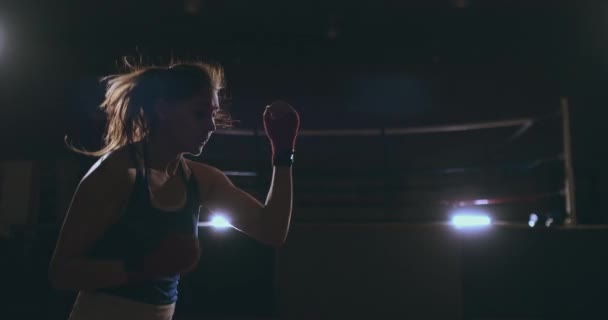 Steadicam-Aufnahmen professionelle schöne Boxerin lädt Schläge während der Durchführung eines Schattenkampfes in einem dunklen Saal in Zeitlupe in einem blauen Kleid und roten Bandagen an ihren Handgelenken. fliegen rund um die — Stockvideo