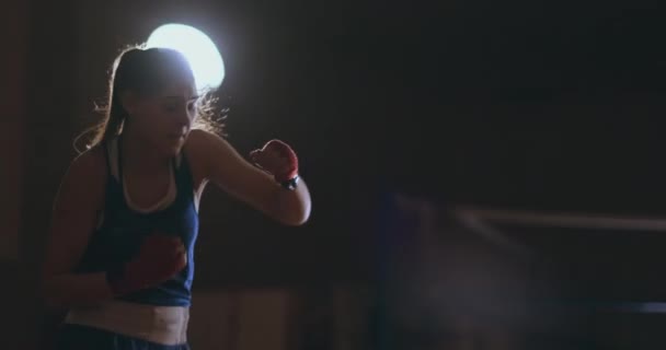 Une belle boxeuse professionnelle se lance dans un combat d'ombre dans une salle sombre au ralenti en vêtements bleus et bandages rouges sur ses poignets. steadicam shot — Video