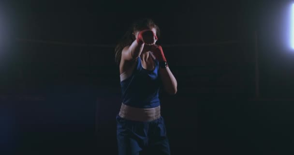 Mirando a la cámara, una hermosa boxeadora golpea contra un fondo oscuro con una luz retroiluminada. Steadicam tiro — Vídeo de stock